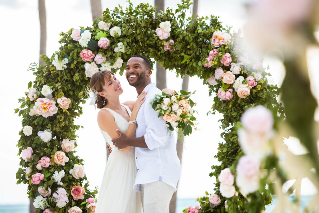 Interracial couple dressed in beige colors getting married in an arch of flowers and greenery