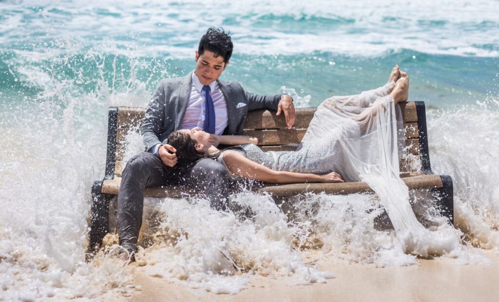 a wedding couple on a beach in the ocean getting splashed by a big wave