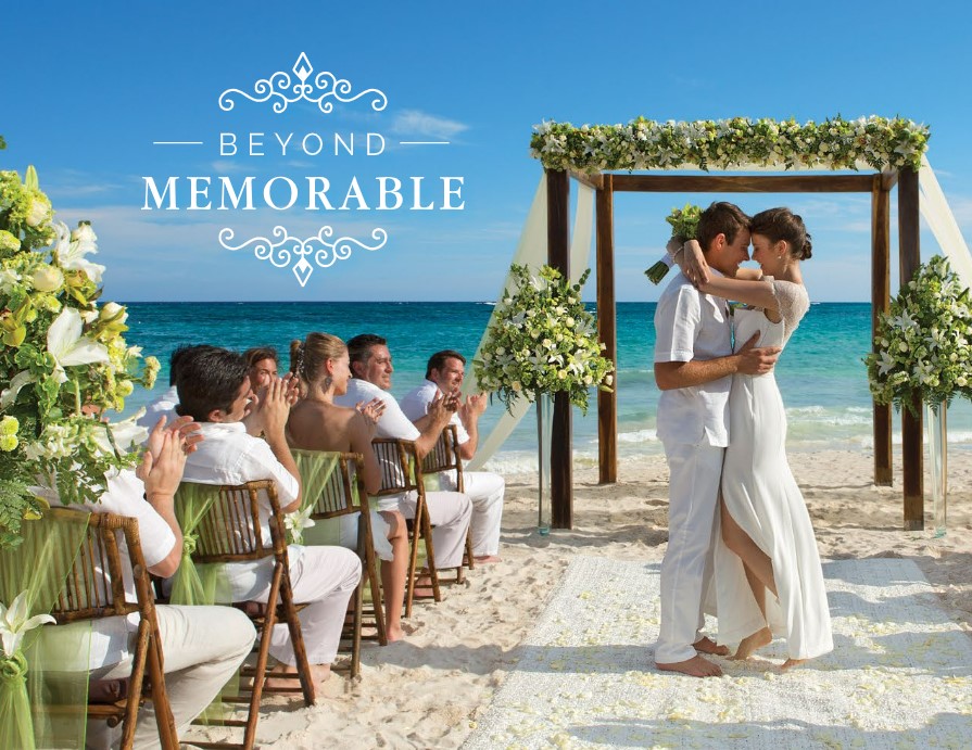 Just married couple holding each other in front of the oceanfront gazebo, all dressed in white with white and green flowers