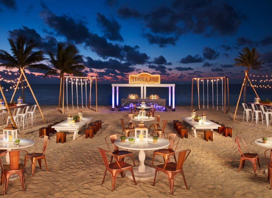 Beach set for a dinner party with brass chairs and white tables, during sunset