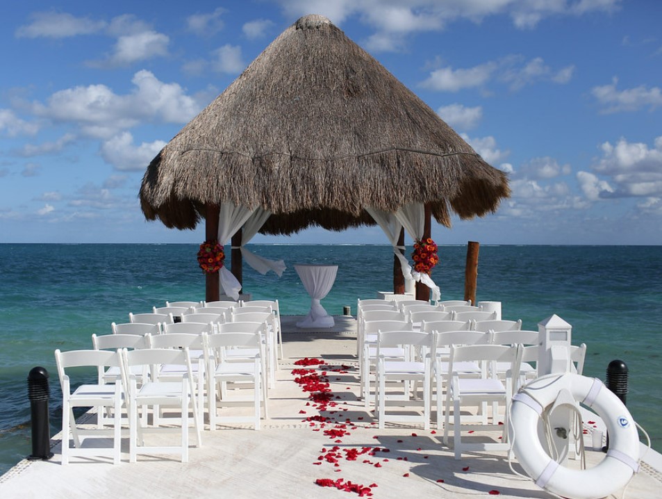 Concrete pier with a roof palapa and white chairs and rose petals set for a wedding
