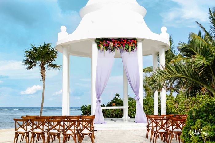 Oceanfront white wedding gazebo with purple drapes, wooden chairs and tropical flowers