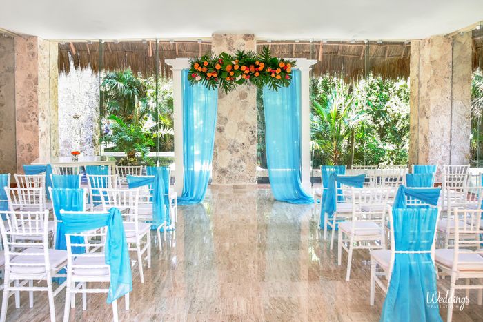 Indoor salon set up for a wedding with a white pergola with blue drapes, white chairs and tropical flowers