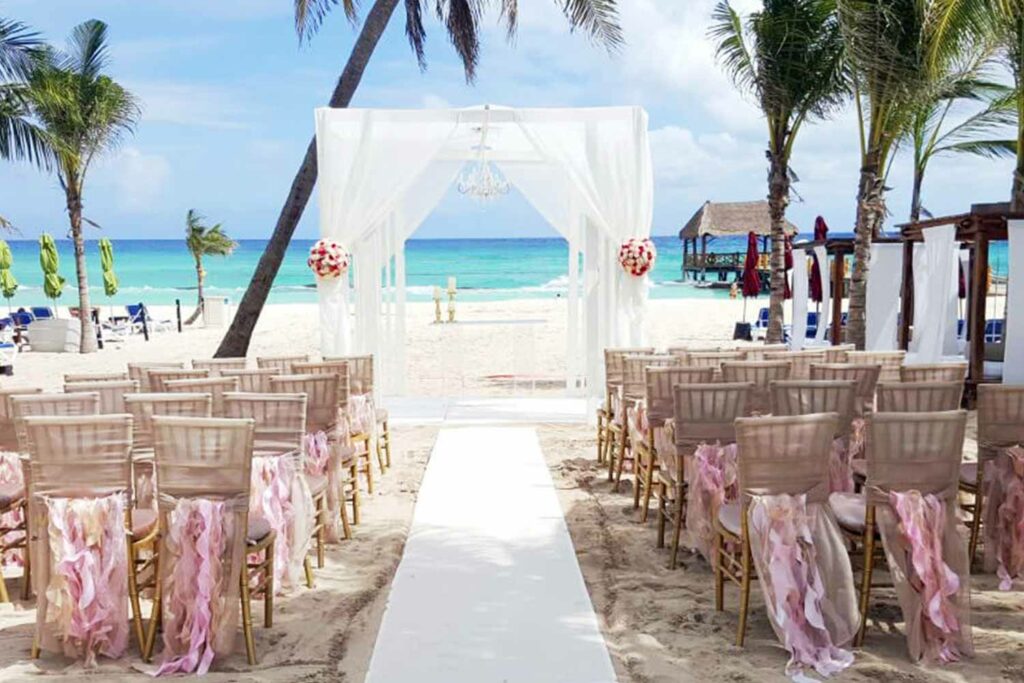 Beach wedding set up with a white pergola and gold chairs with nude ribbons