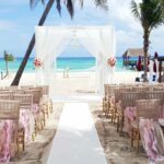 Beach wedding set up with a white pergola and gold chairs with nude ribbons