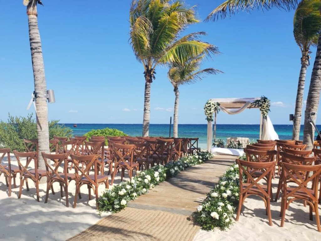 Beach wedding pergola with white drapes, wooden chairs and white flowers with green leaves