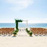 Beach wedding set up with a white pergola with greenery and wooden chairs