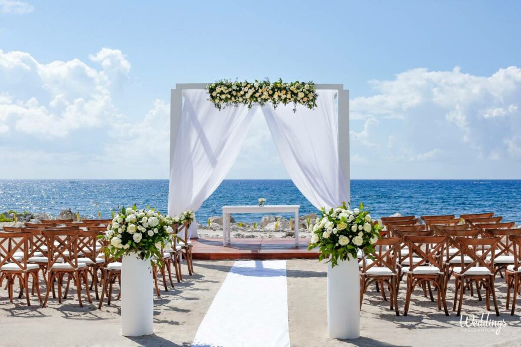 Oceanfront wedding pergola with white drapes, flowers and wooden chairs