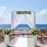 Oceanfront wedding pergola with white drapes, flowers and wooden chairs