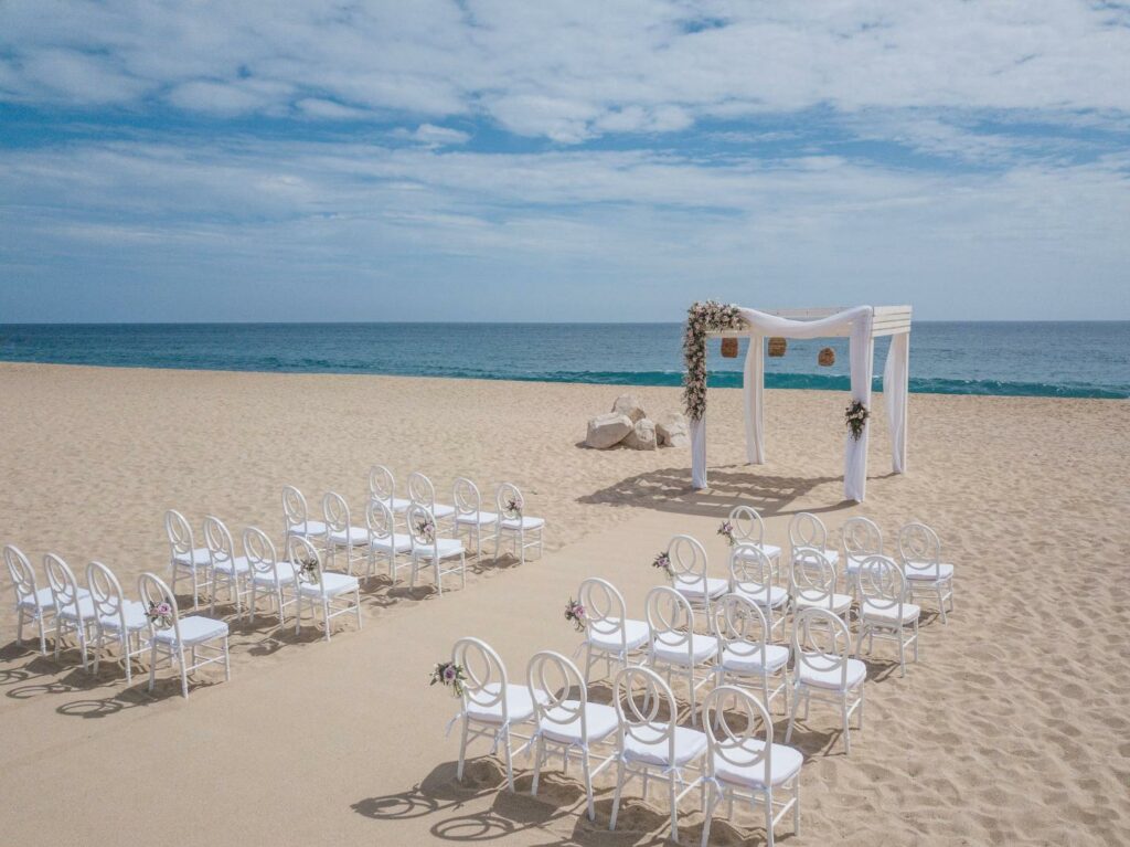 wedding ceremony set up at a beach in los cabos with a white pergola with drapes and flowers and white chairs
