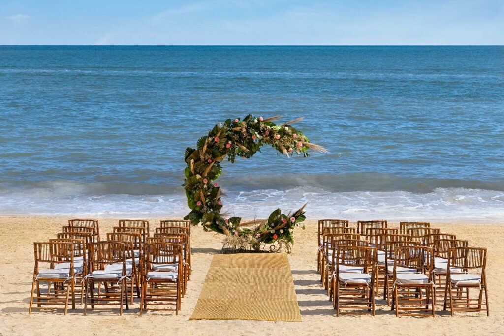 Beach wedding ceremony set up with an arch with flowers and wooden chairs