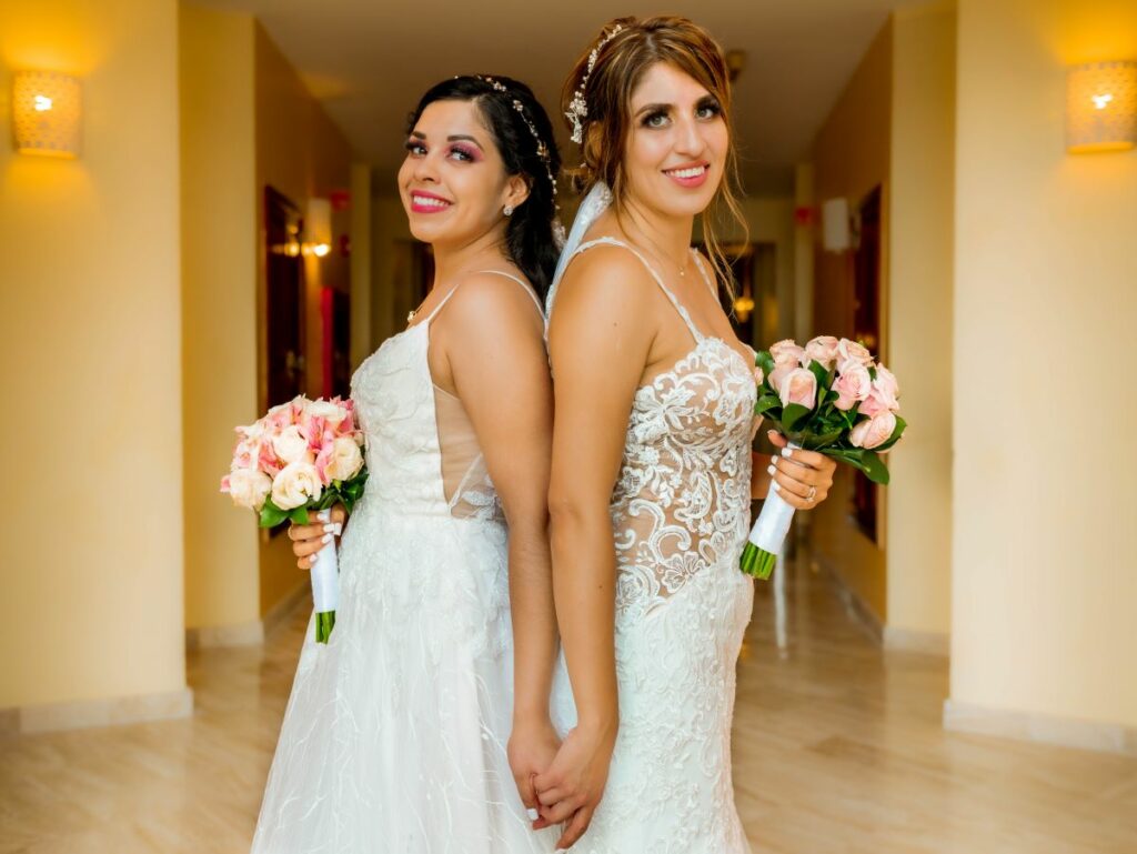 Two brides dressed in white dresses holding hands and a rose bouquet