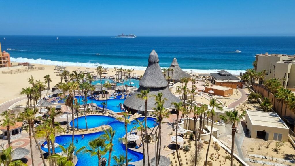 Aerial view of a resort in Los Cabos, with pools, palm trees and the ocean at the back with a large cruiser ship