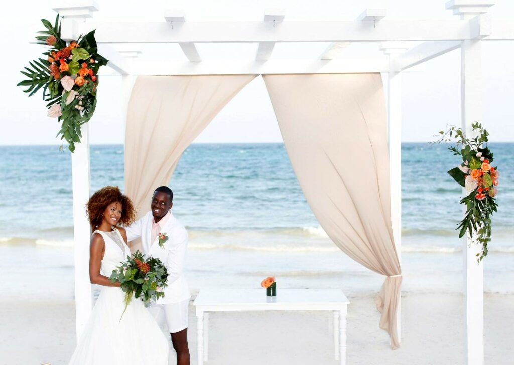 wedding couple posing below an ocean front pergola with tropical flowers and beige drapes