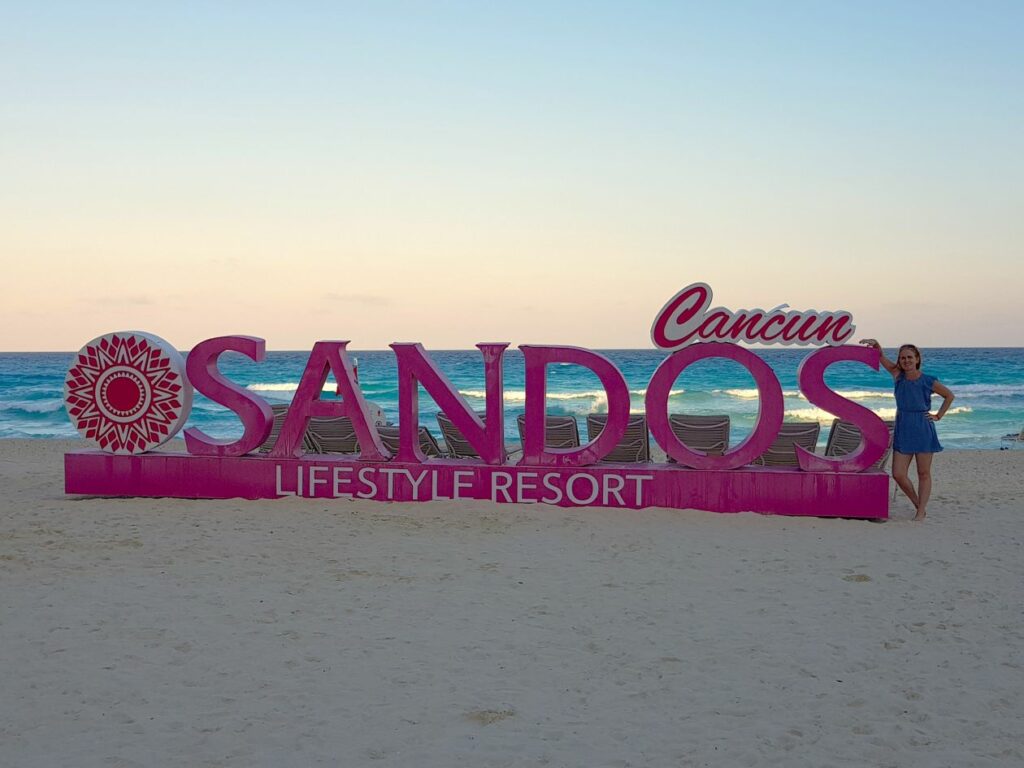 Lisa wright from hola weddings standing next to sandos cancun sign on the beach