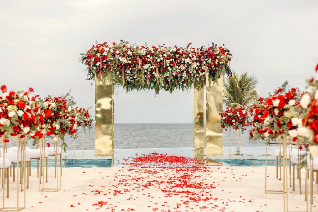wedding ceremony set up on a platform over the water, with a golden structure with lots of red and white roses, rose petals on the aisle and also flower arrangements