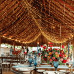 A wedding set up under a palapa roof decorated with string lights and tables with blue glasses, and colorful floral arrangments