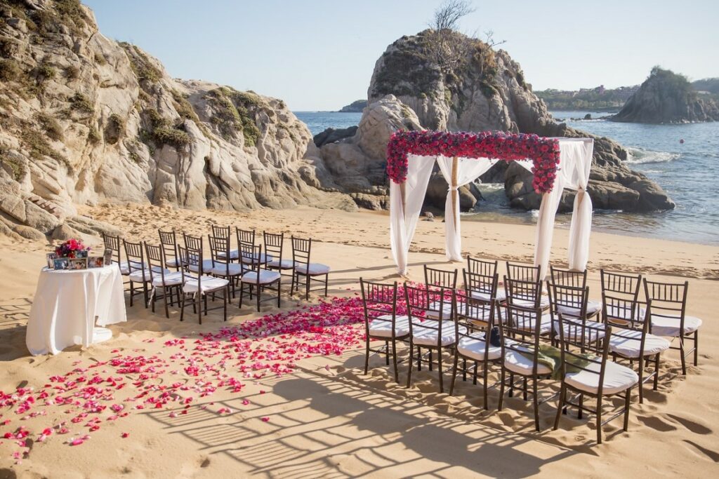 A beautiful wedding ceremony set up on the beach, with rocks in the background and red rose petals on the aisle